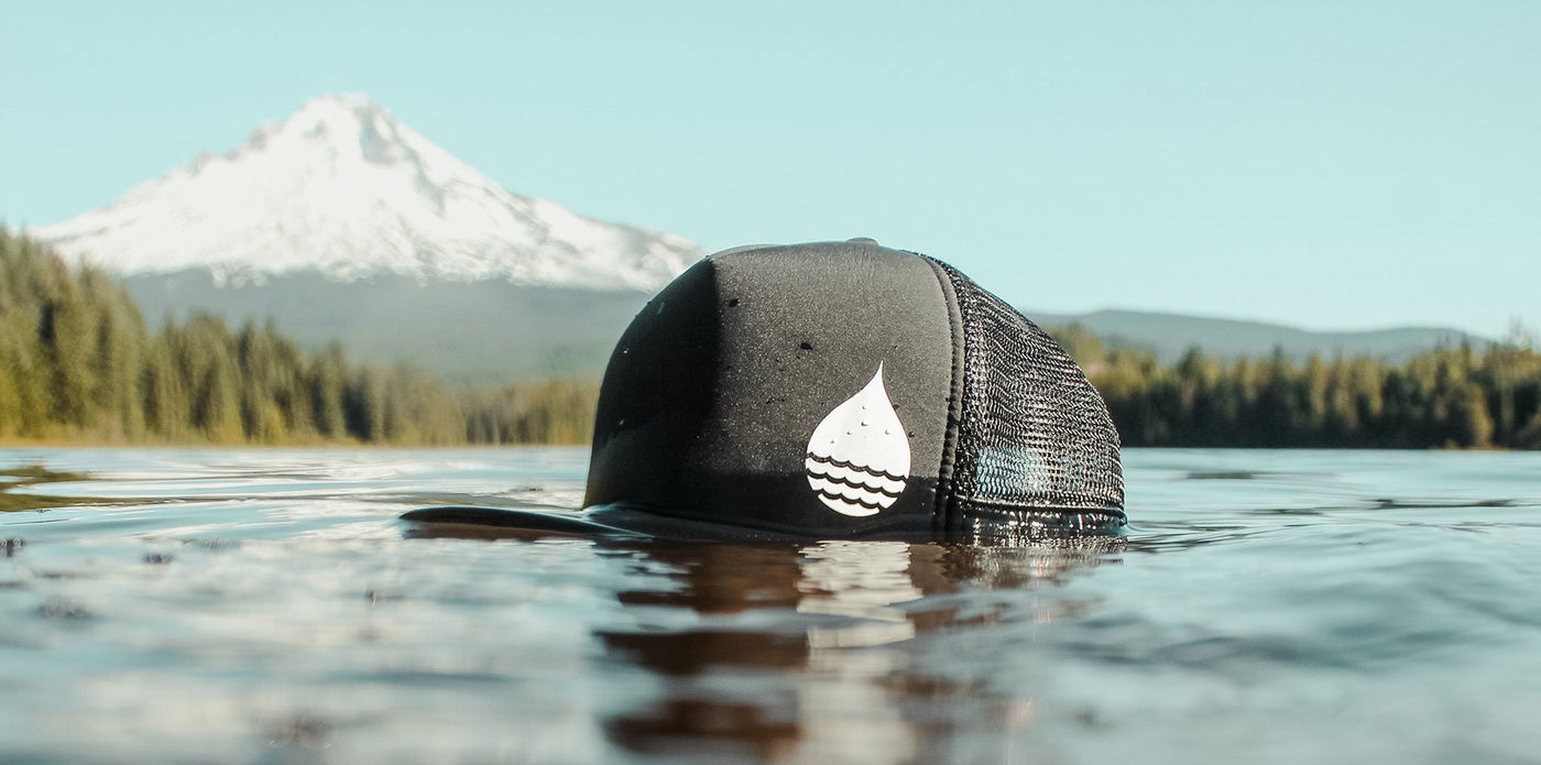The BUOY WEAR black floating trucker hat in the lake with a view of Mount Hood, Oregon in the background.